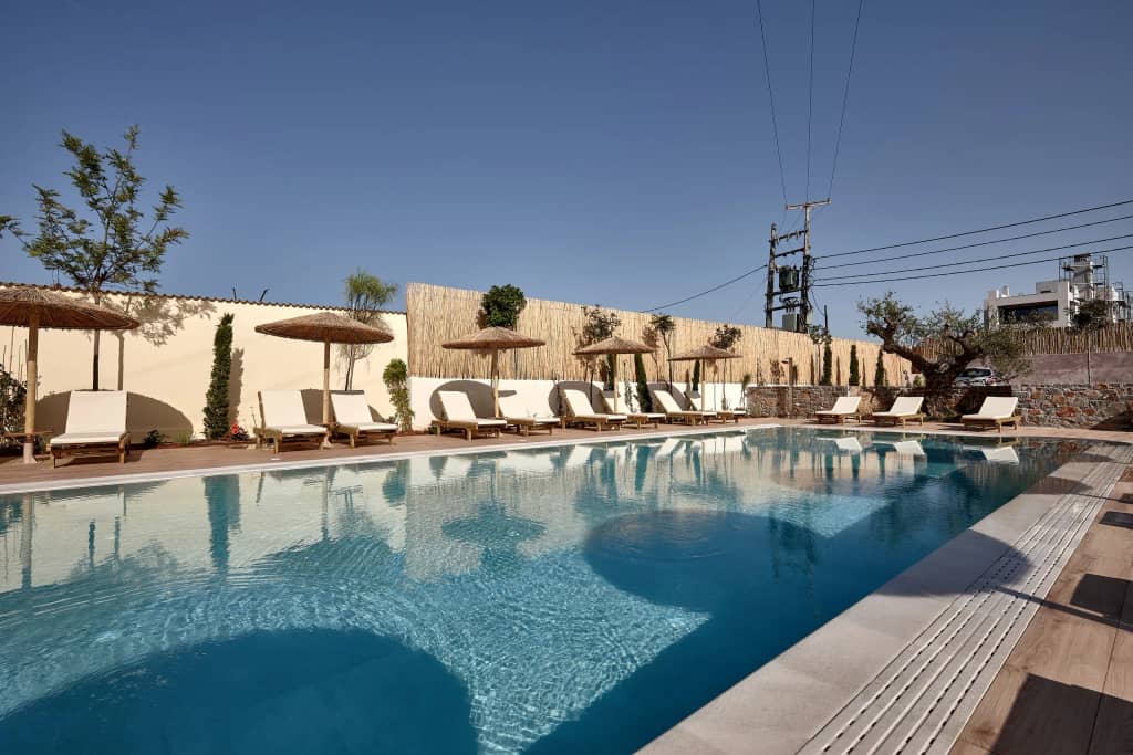 Sparkling blue pool edged by loungers, straw umbrellas and a mature olive tree under a clear sky